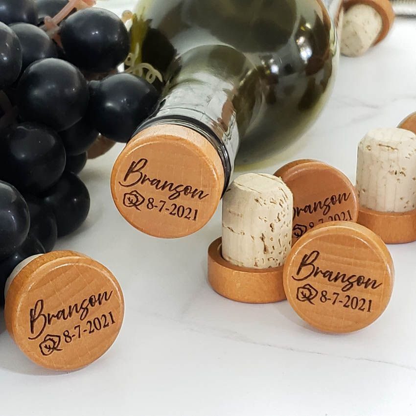 Wooden wine stoppers engraved with a name and date next to purple grapes.
