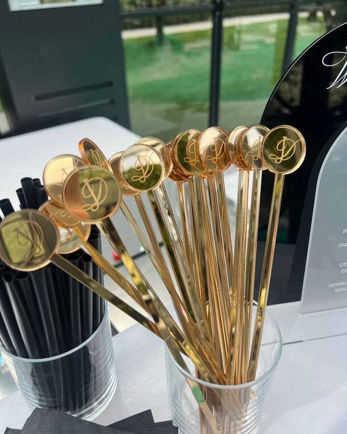 Gold cocktail stirrers with engraved letter D in a clear glass holder.