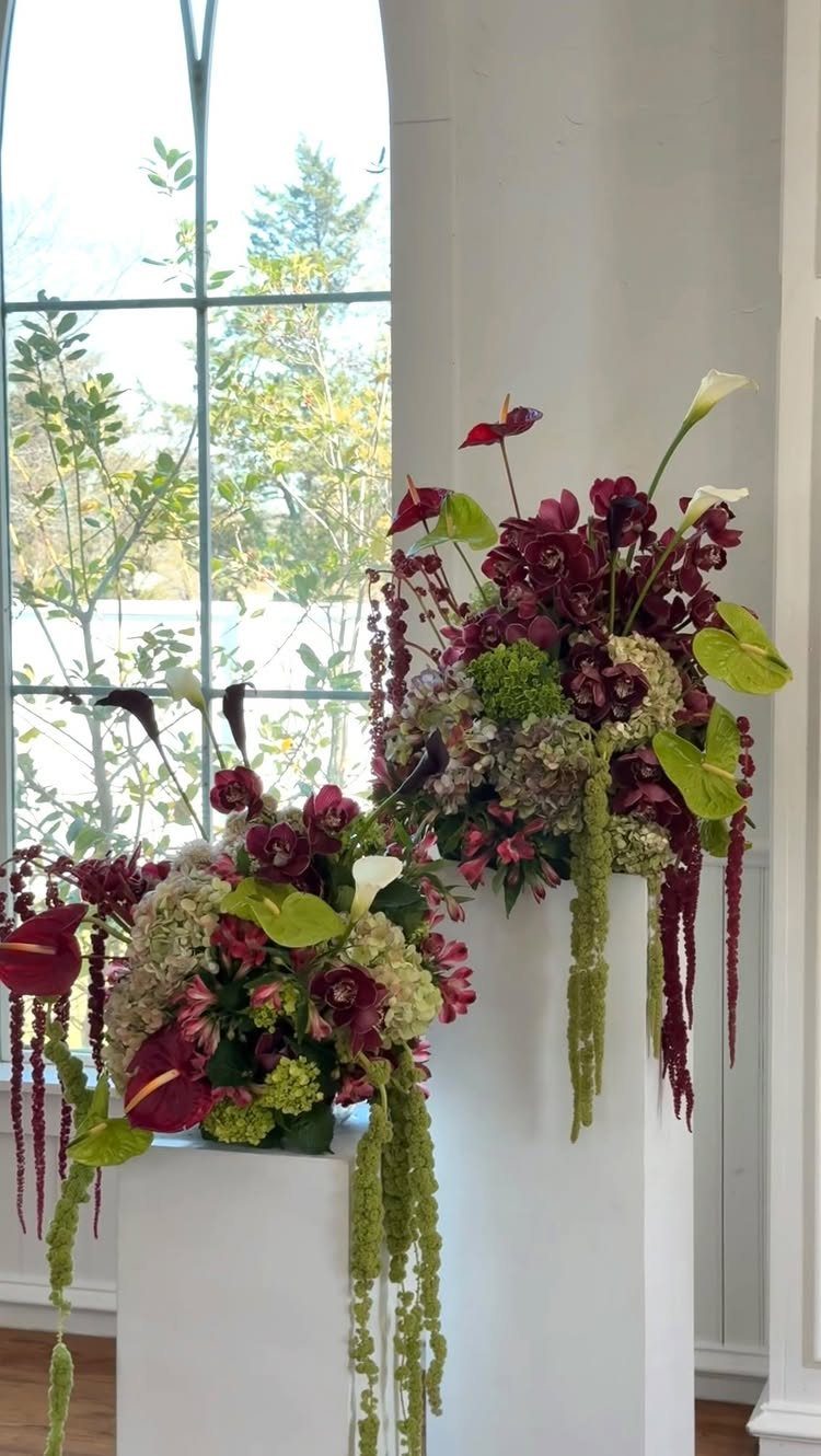 Burgundy and green floral arrangements on white pedestals by a bright window.