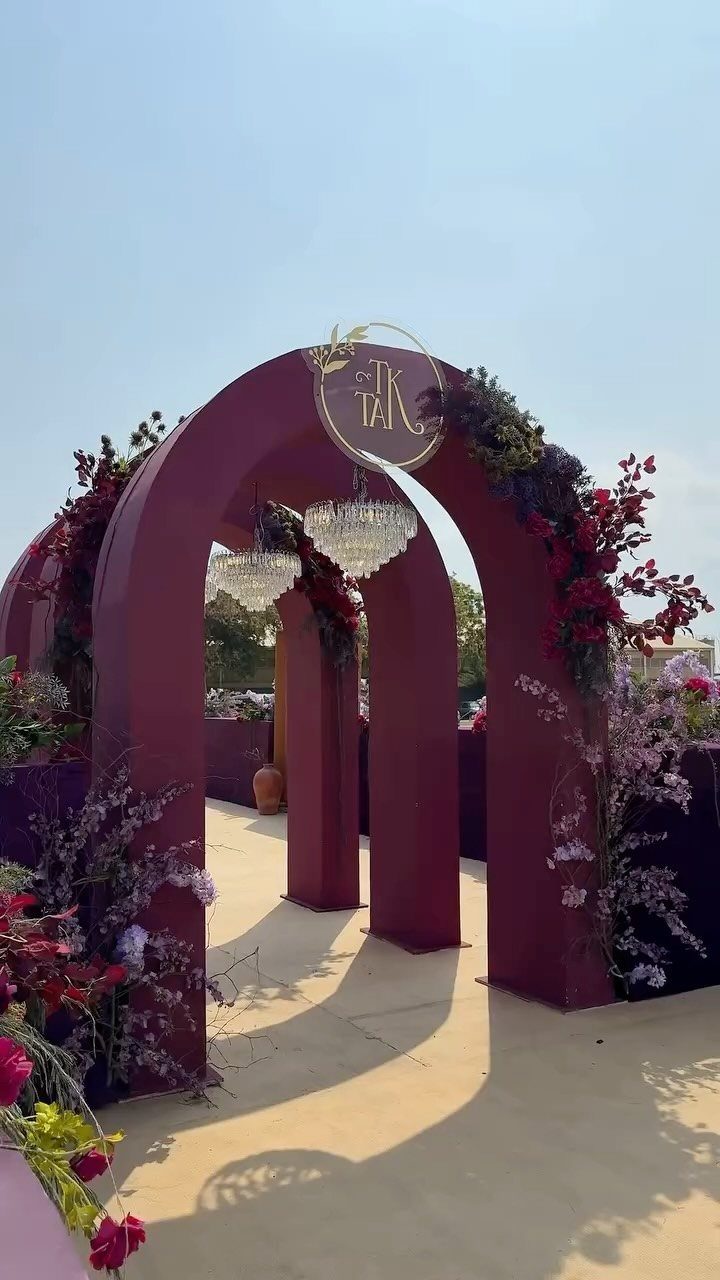 Deep plum arched wedding structures decorated with red flowers and crystal chandeliers outdoors.