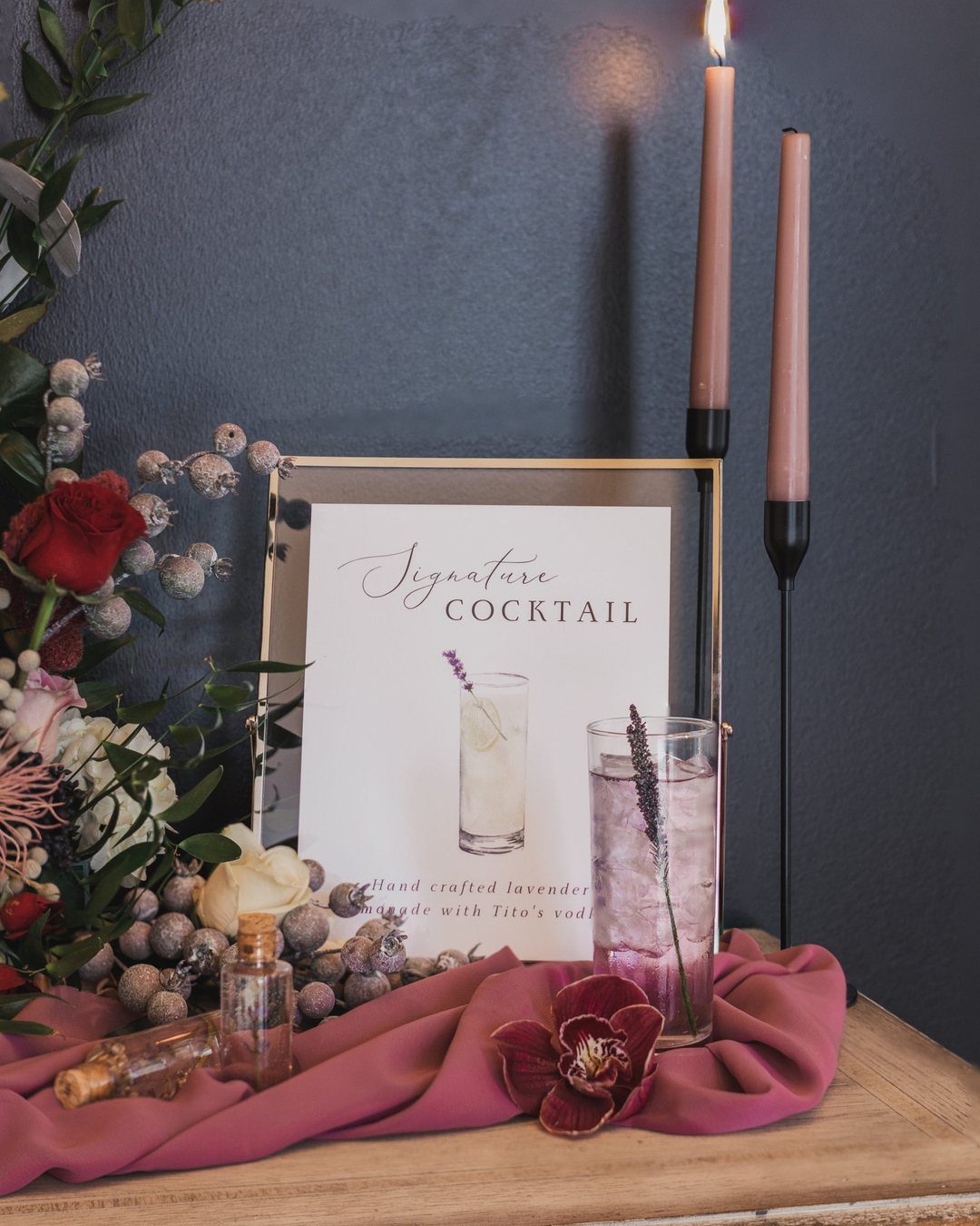 Mauve wedding cocktail setup with candles, floral arrangement, and a framed signature drink sign.
