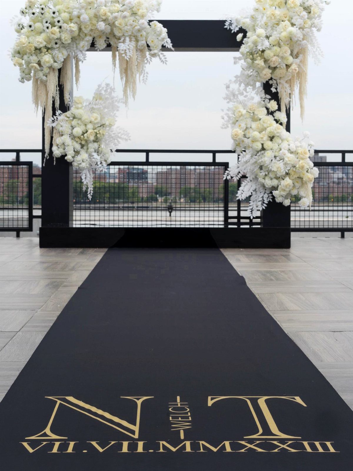 Black aisle runner with gold text leading to a white floral wedding arch.
