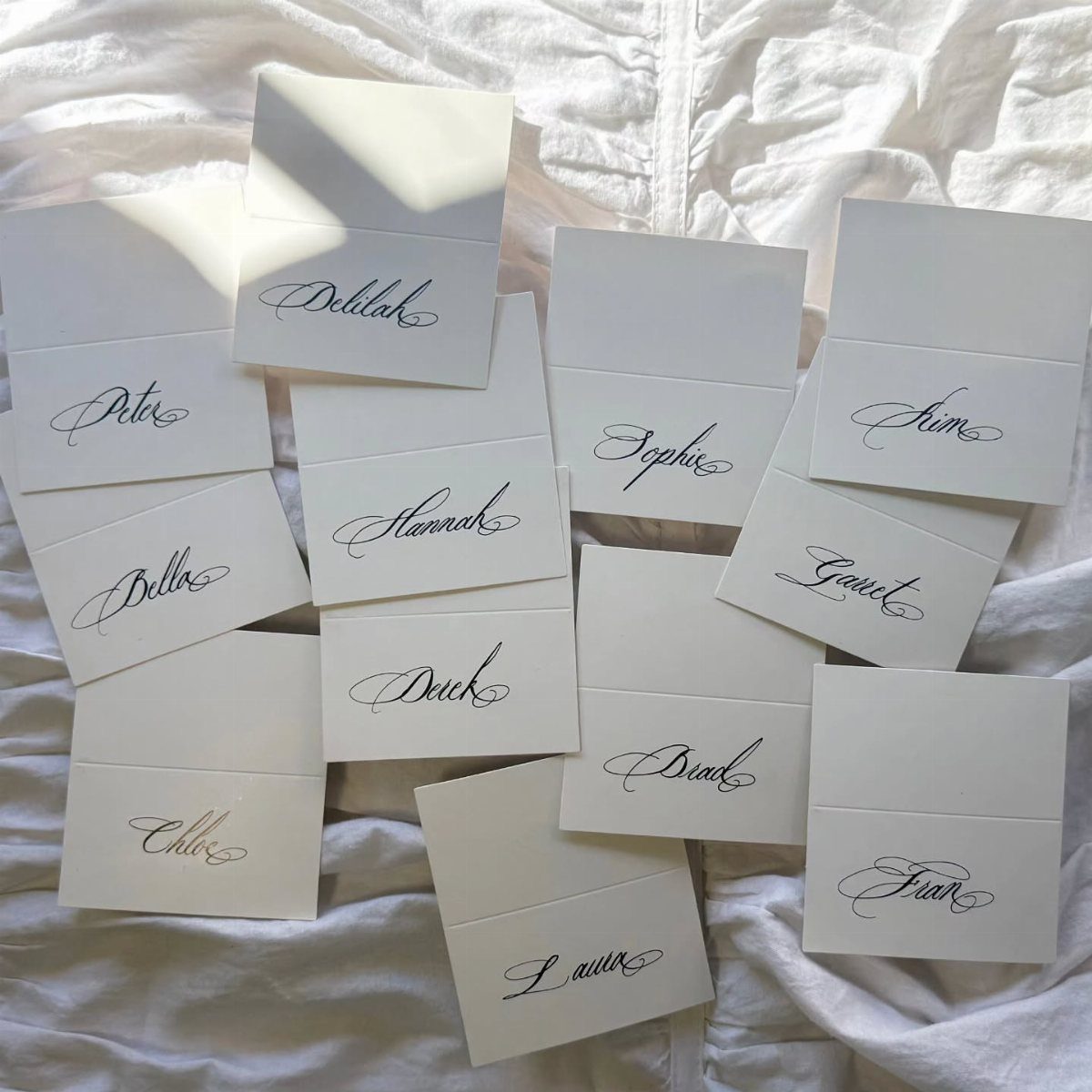 White place cards with black cursive calligraphy names scattered on white fabric.