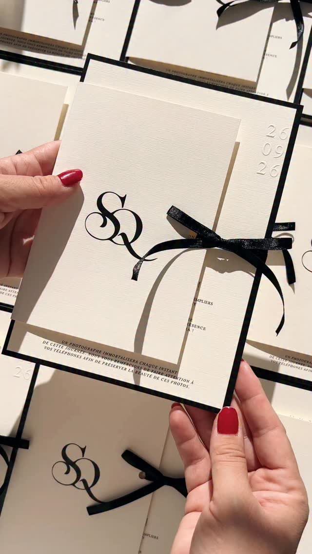 White wedding invitation with black monogram and black ribbon held by hands.