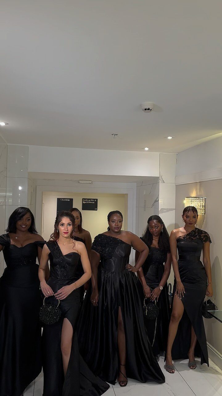 Six women in formal black evening gowns posing together in a white room.