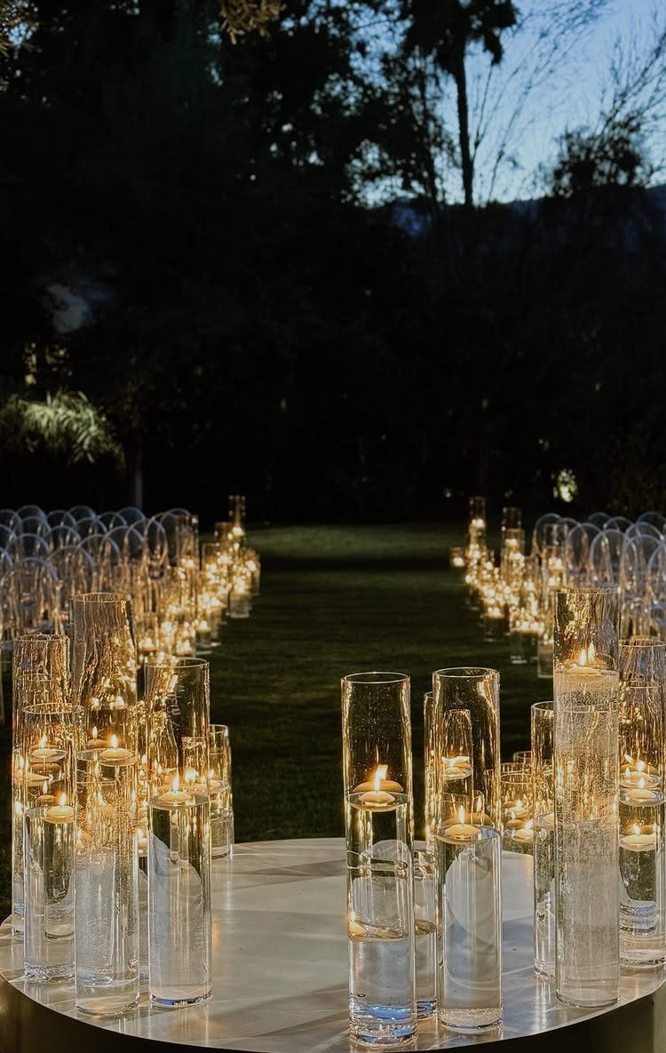 Floating white candles in glass cylinders lining a grassy outdoor wedding aisle at dusk.