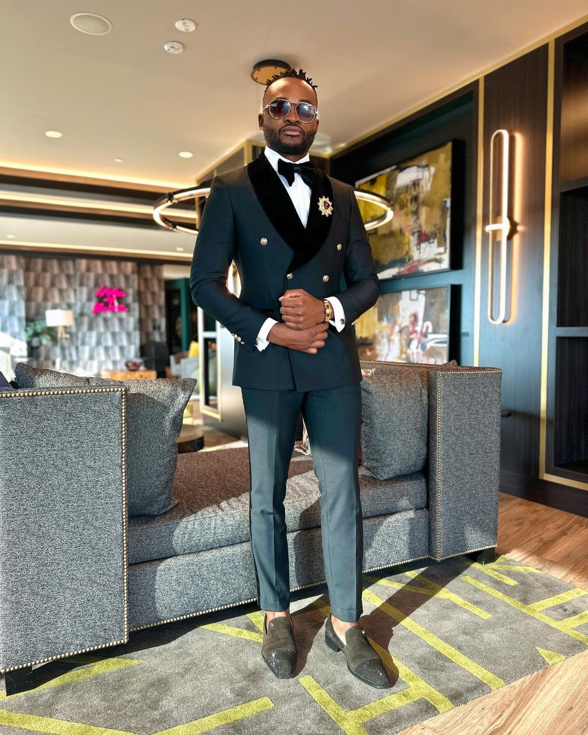 Man in a black double-breasted tuxedo and bow tie standing in a modern lounge.