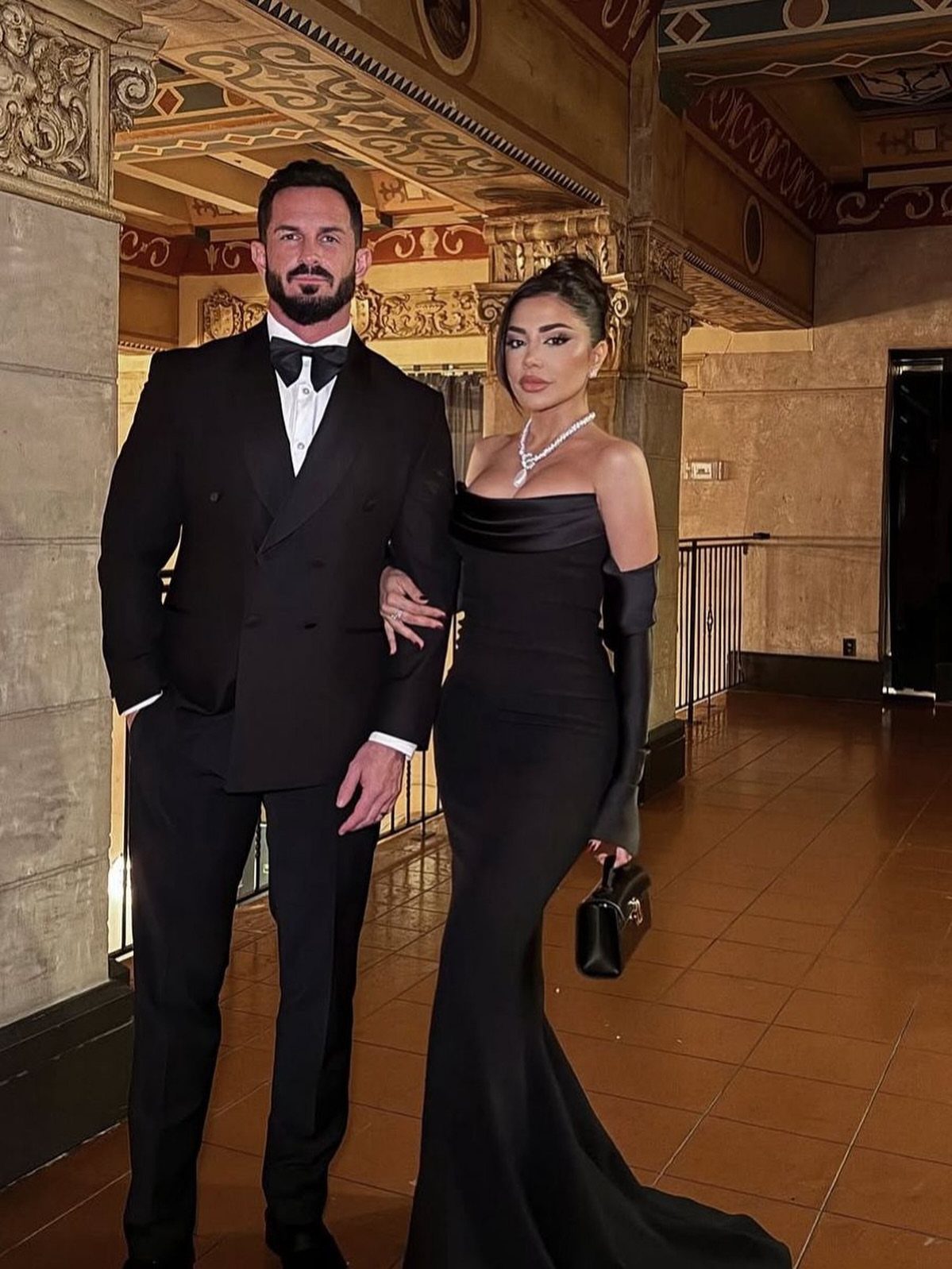 Couple in formal black tie attire posing in an ornate classical hallway.