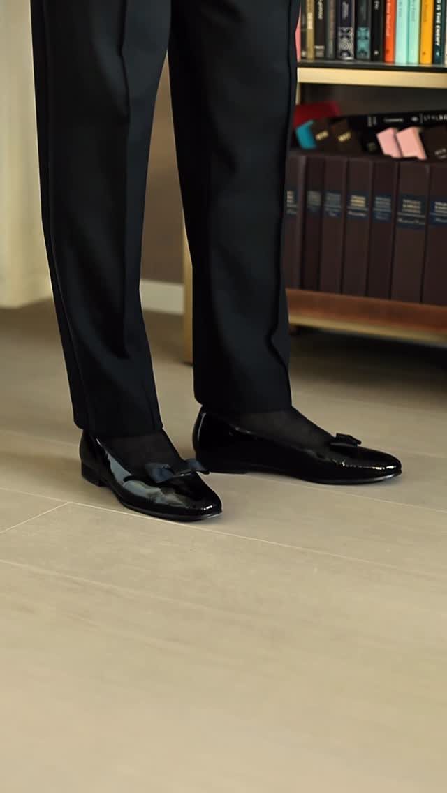 Black formal trousers and patent leather loafers with bows on a wooden floor.