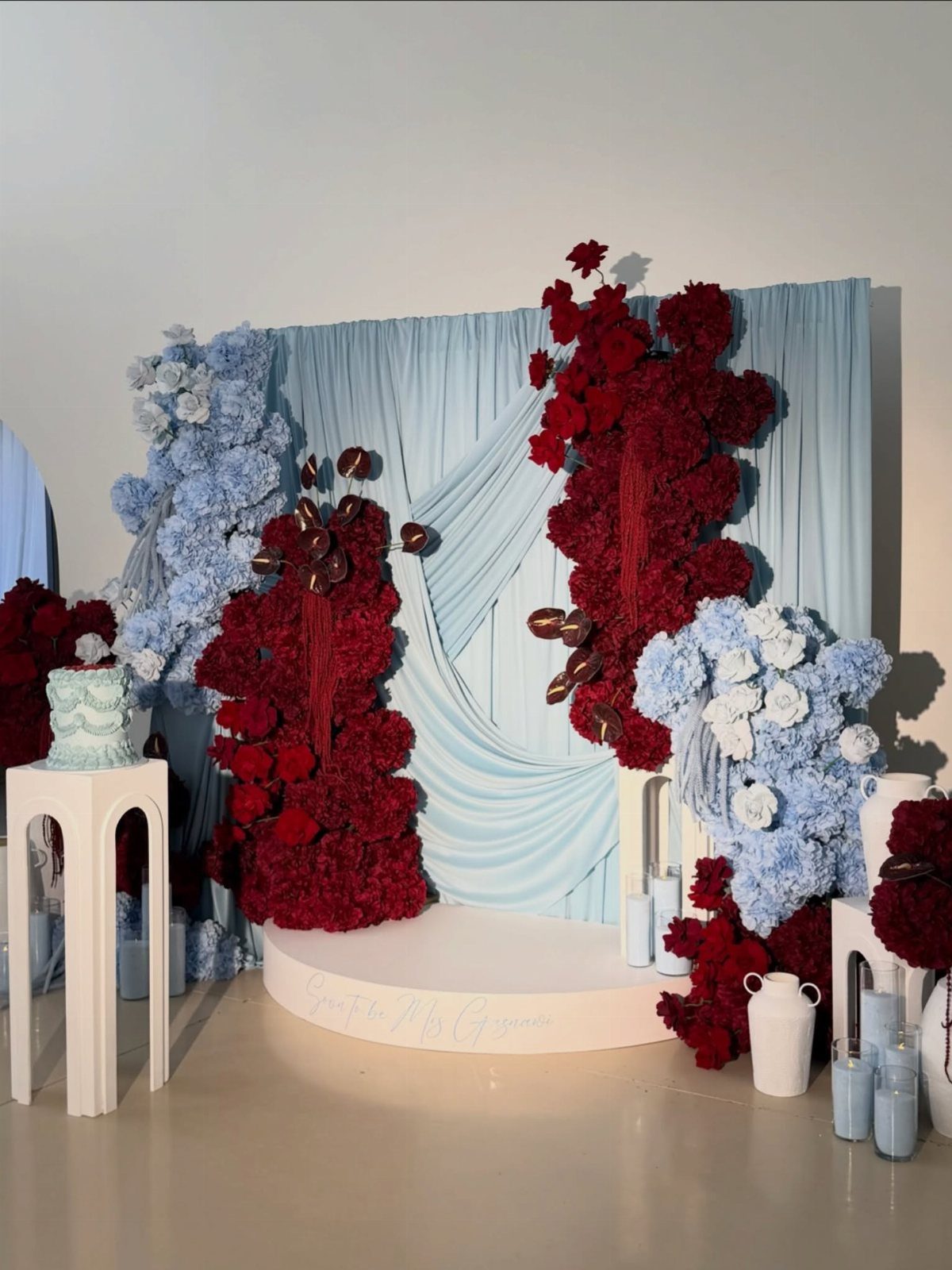 Wedding backdrop with light blue drapery and large red and blue floral arrangements.