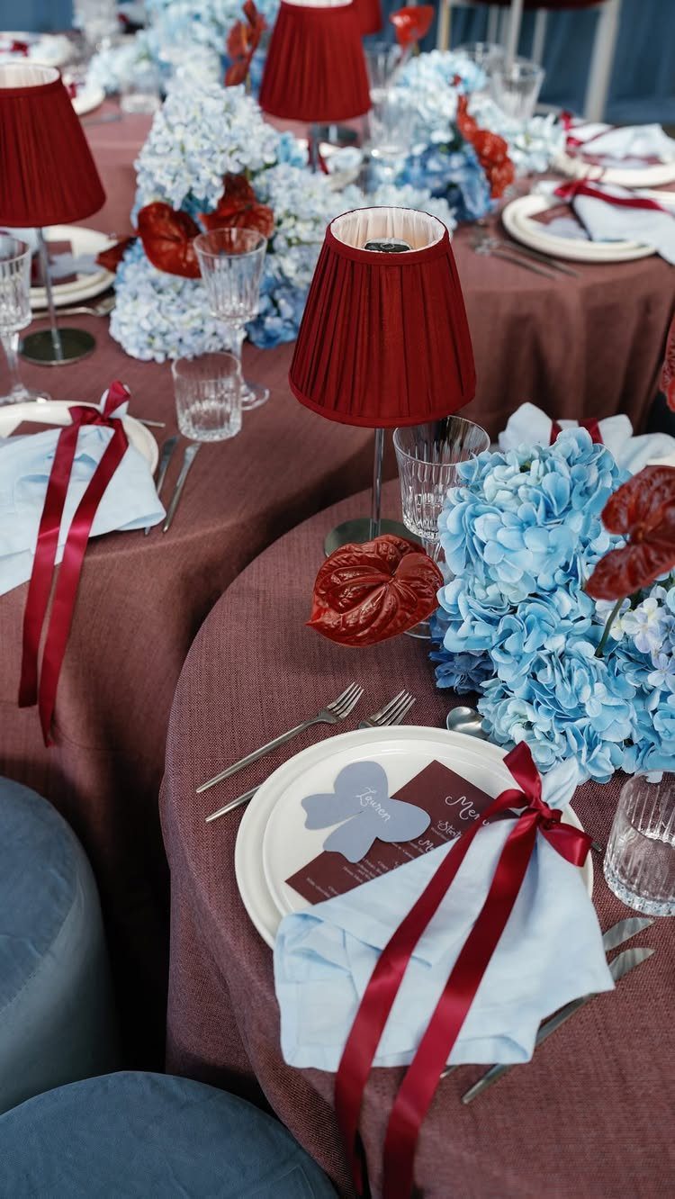 Wedding table setting with burgundy linens, blue hydrangeas, and red lampshades.