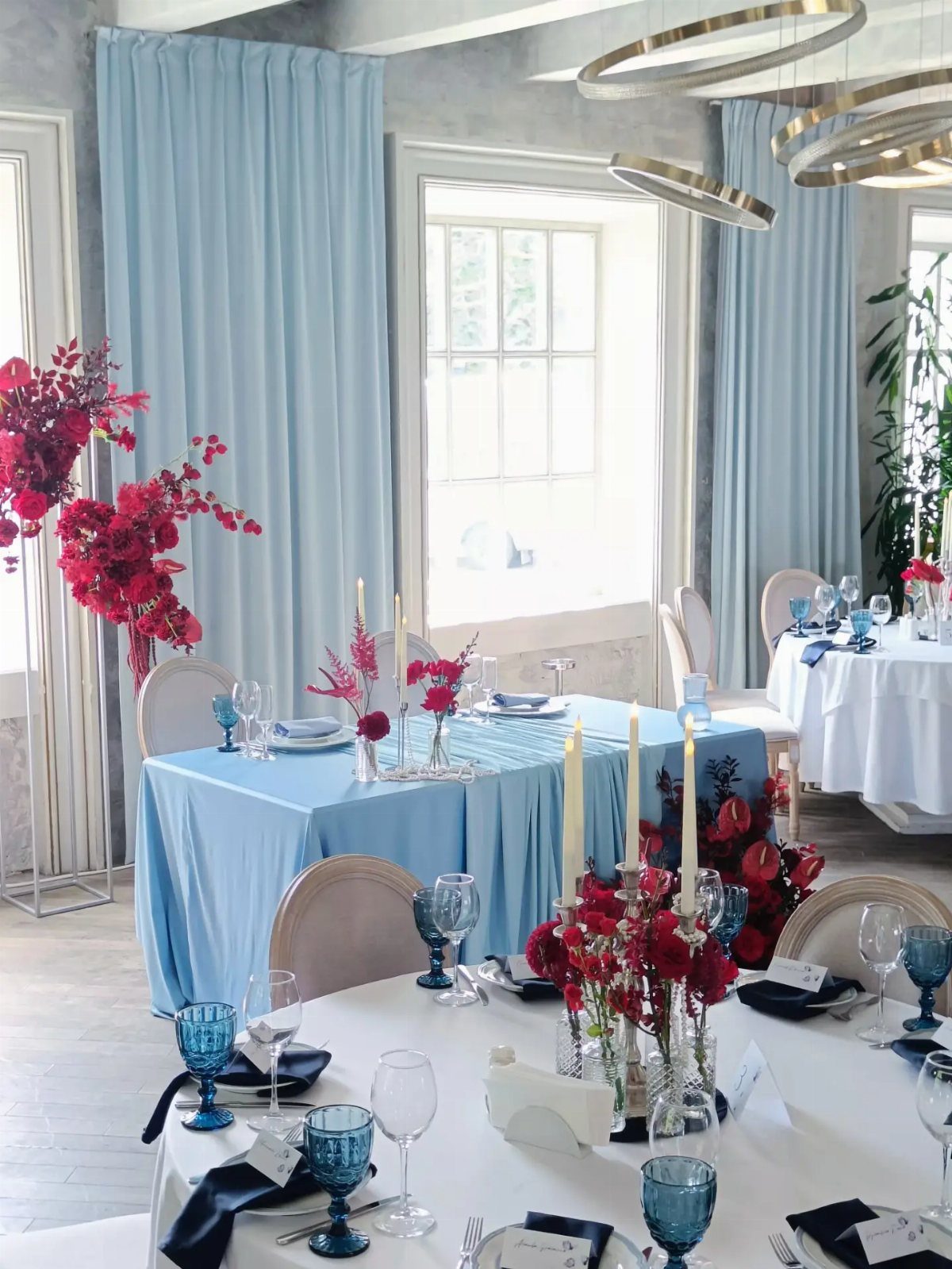 Red flower centerpieces with white candles on blue and white wedding tables.