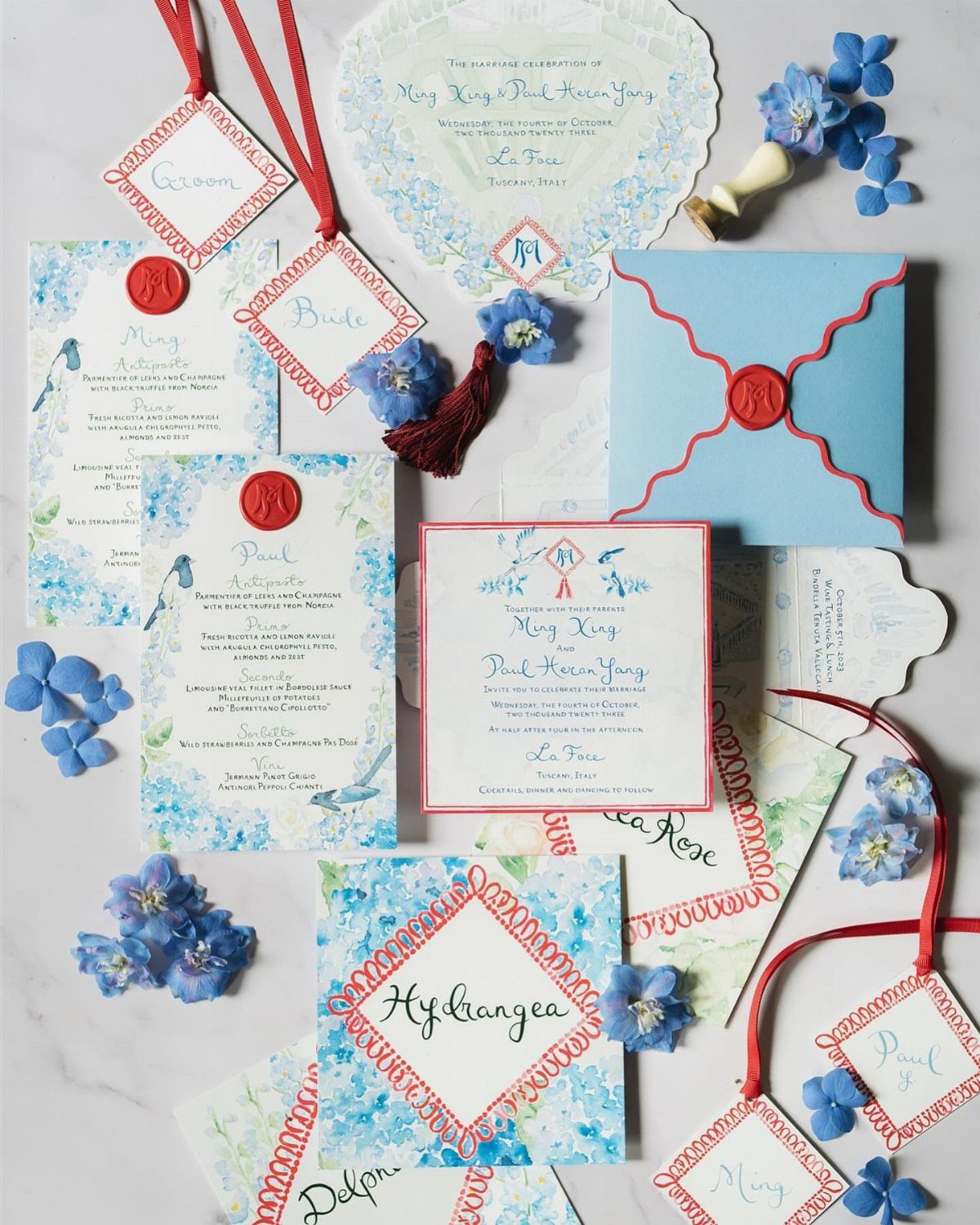 Wedding invitations and stationery with blue floral designs and red accents on marble.