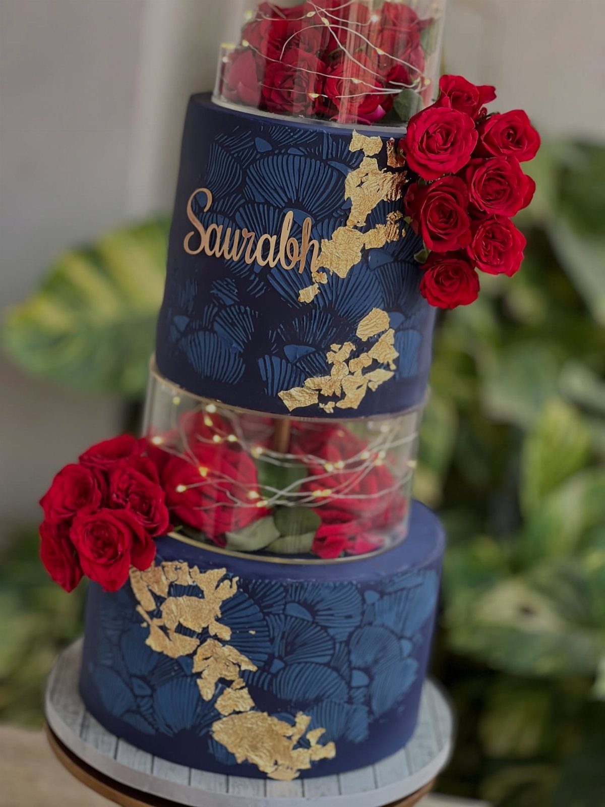 Navy blue and gold tiered cake decorated with red roses and fairy lights.