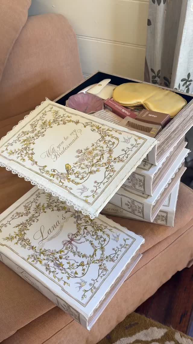 Book-shaped gift boxes with gold floral designs containing bridesmaid favors on a sofa
