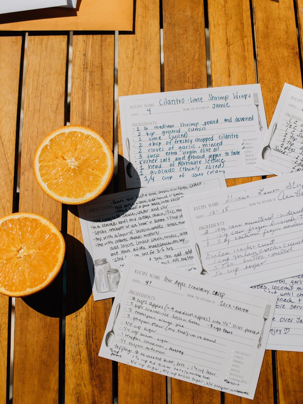 Handwritten recipe cards and orange halves resting on a wooden slatted table.