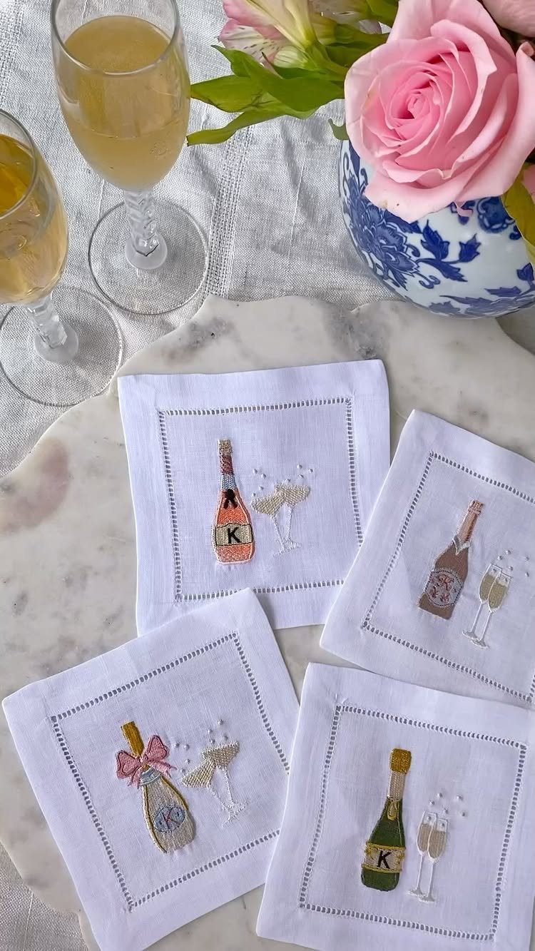 Embroidered white napkins with champagne bottles next to champagne glasses and pink roses.
