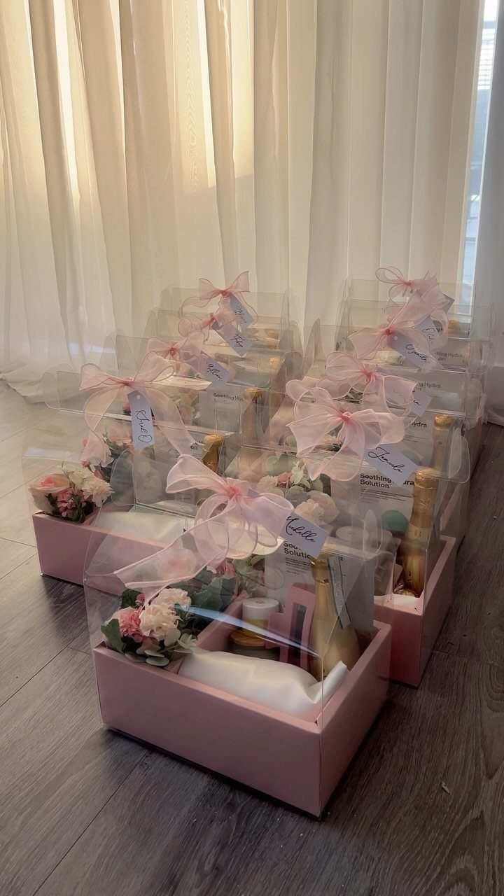 Pink gift boxes with clear lids containing mini champagne bottles and small flowers.