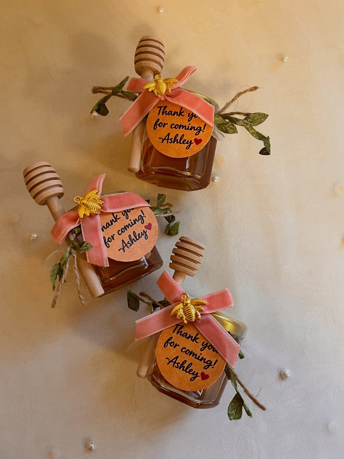 Three mini honey jars with pink ribbons, honey dippers, and personalized thank you tags.