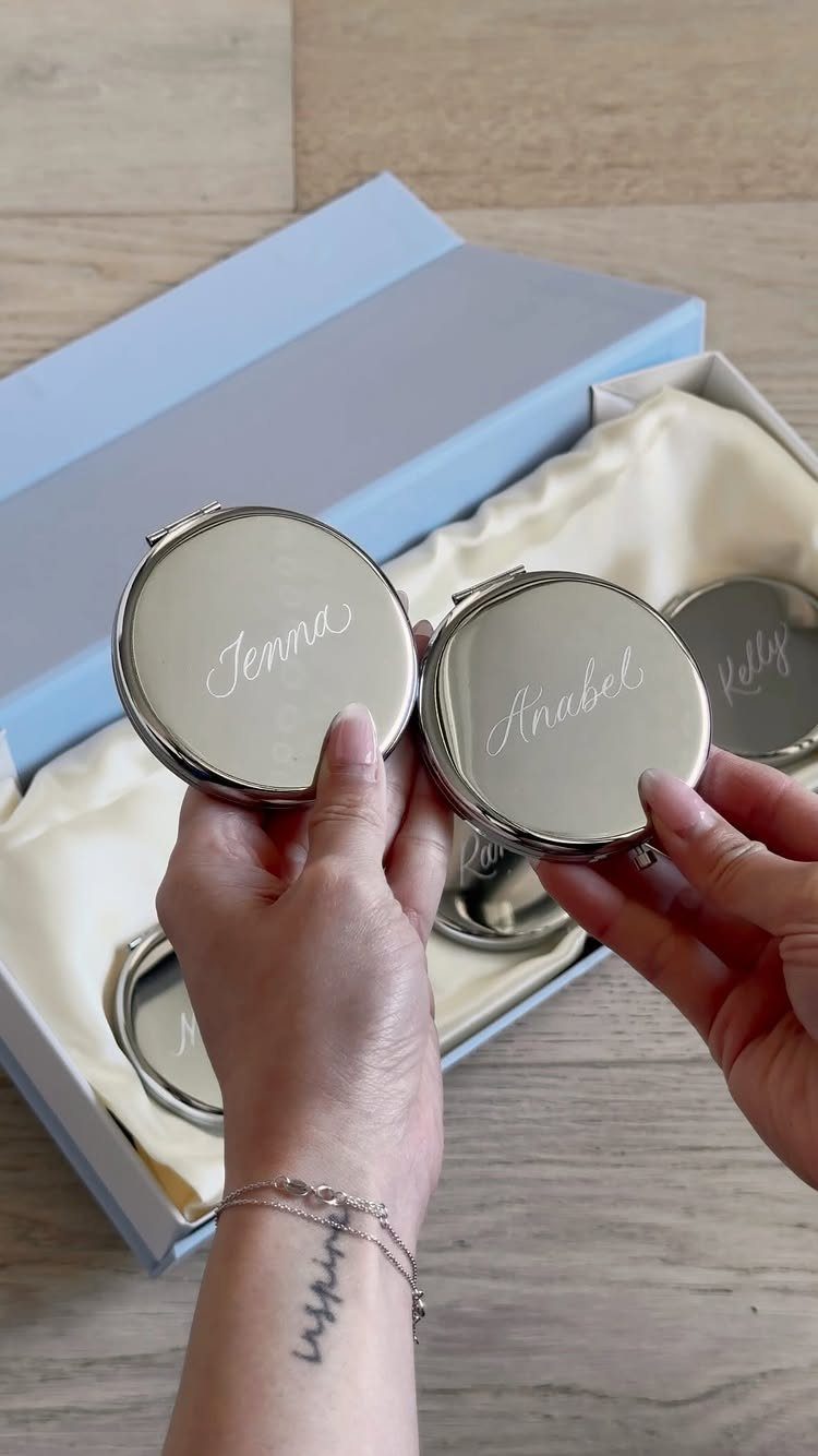 Silver compact mirrors with engraved names held over a light blue gift box.