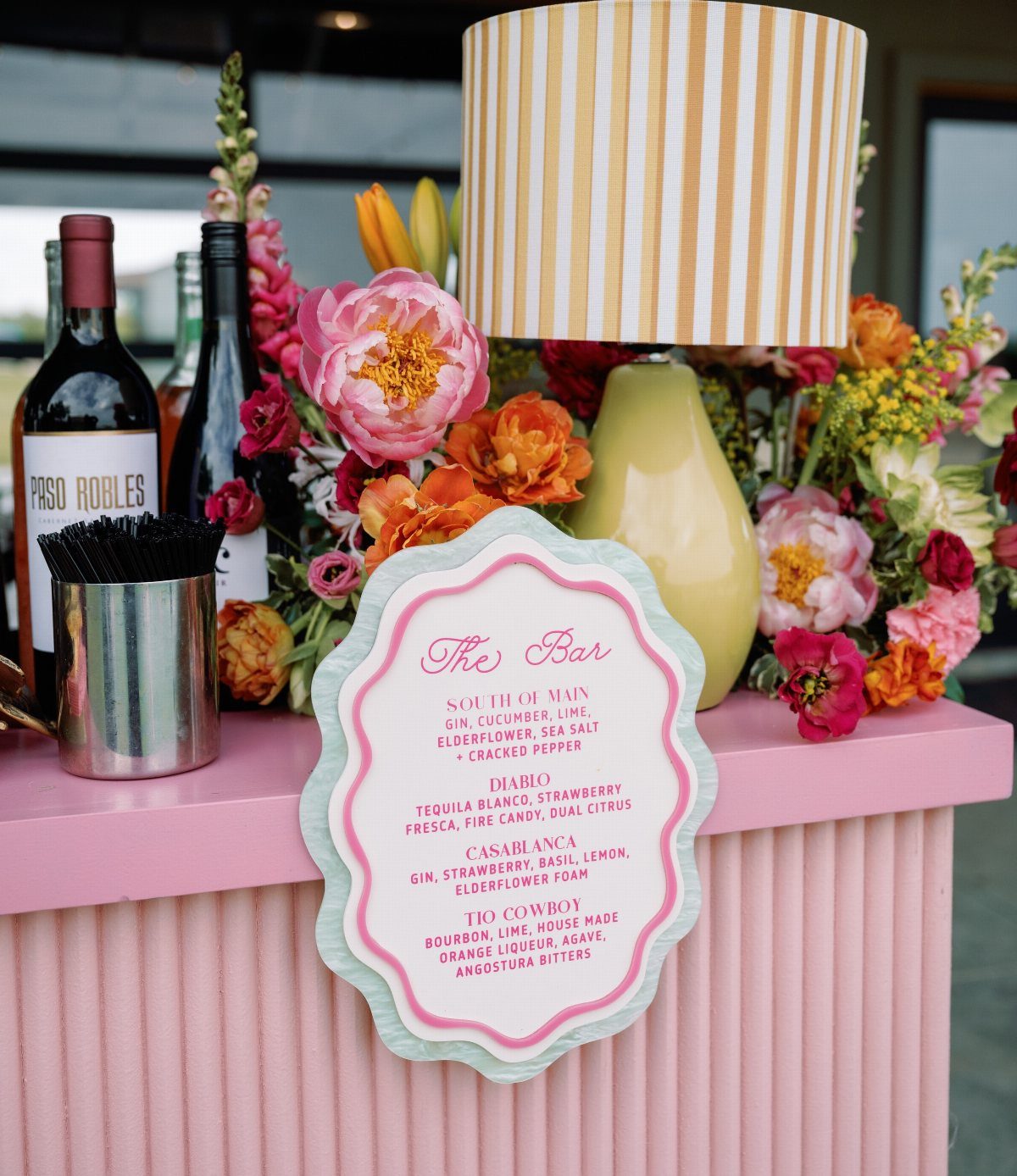 Pink bar counter with colorful floral arrangements, a yellow lamp, and drink menu.