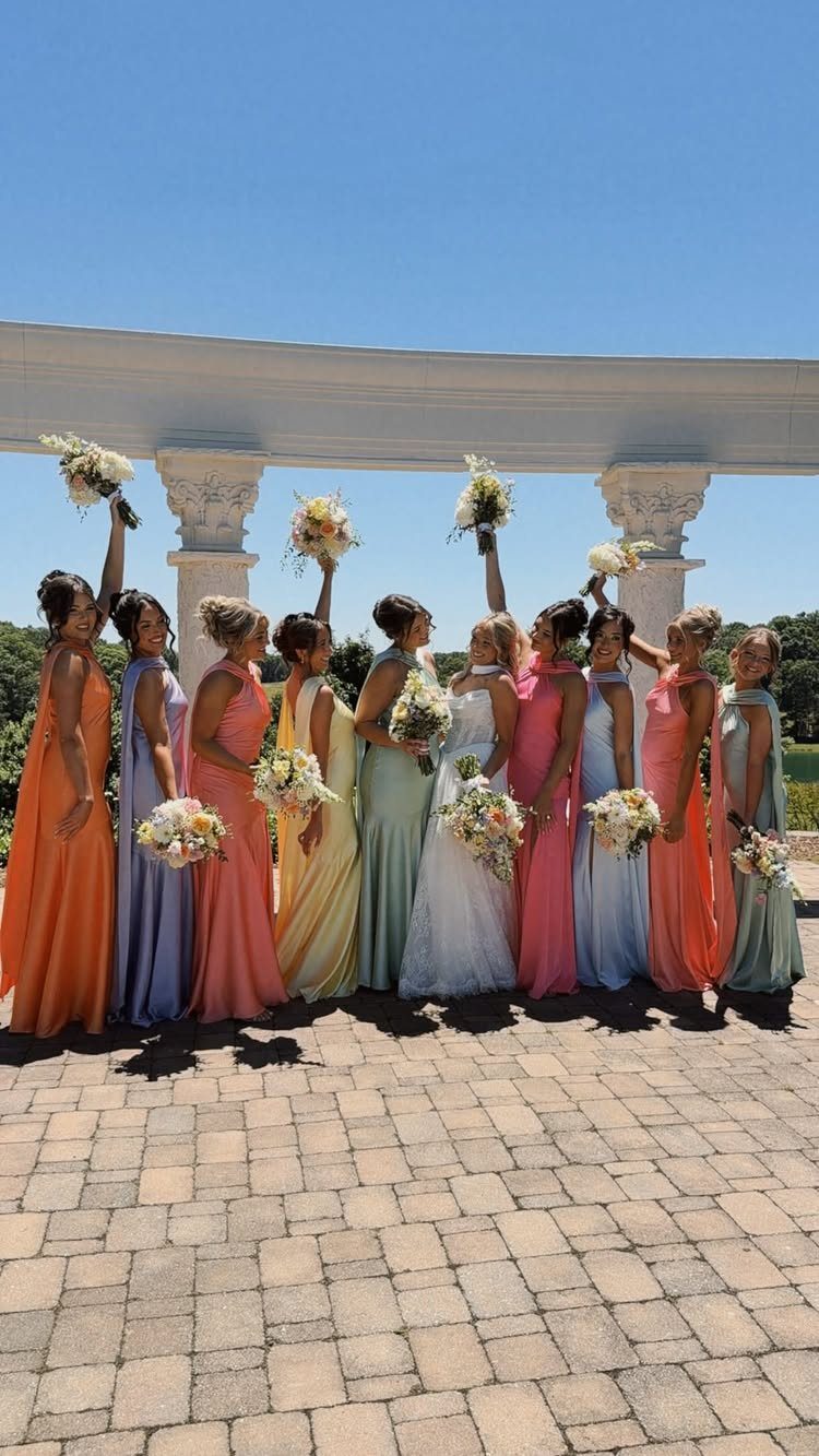 Bride and bridesmaids in mismatched bright colored dresses posing under a white arch.