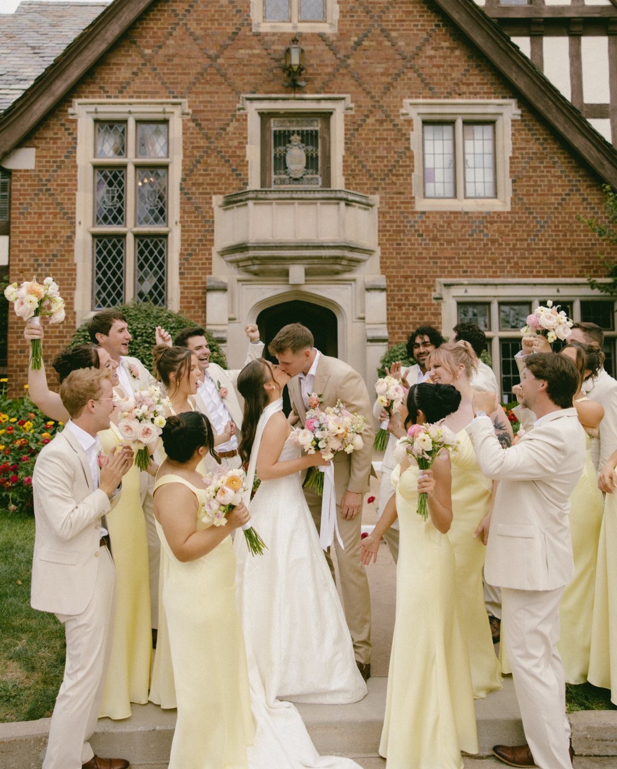 Wedding party in butter yellow and off-white attire posing before a brick manor.