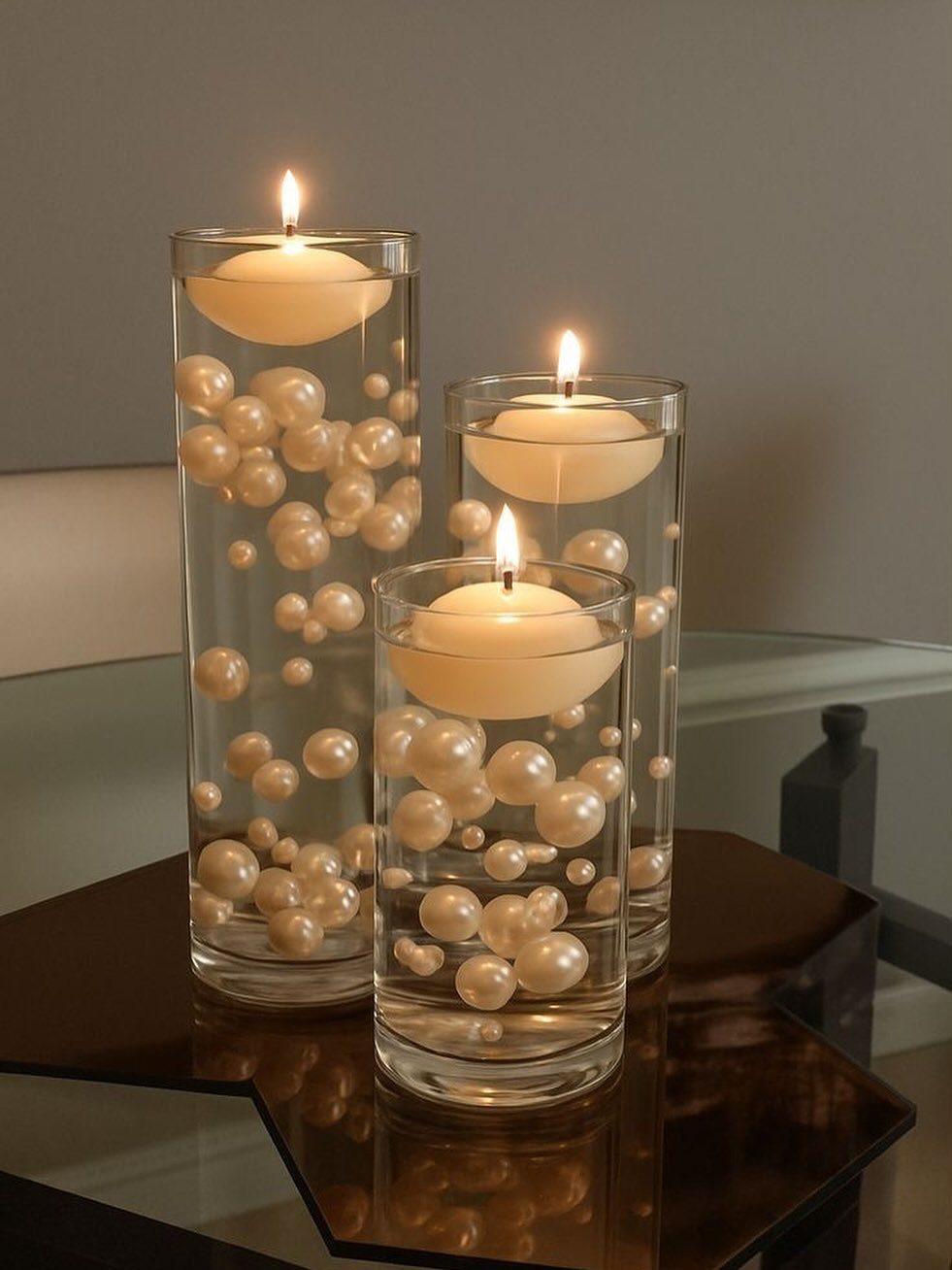 Three glass vases with floating white candles and pearls on a reflective table