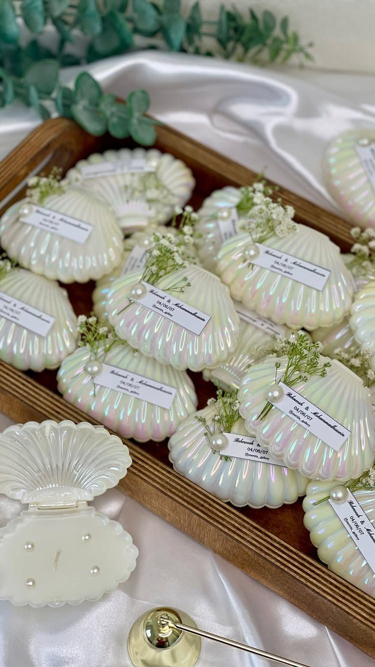 Shell-shaped white iridescent candles with labels in a wooden tray on satin fabric.