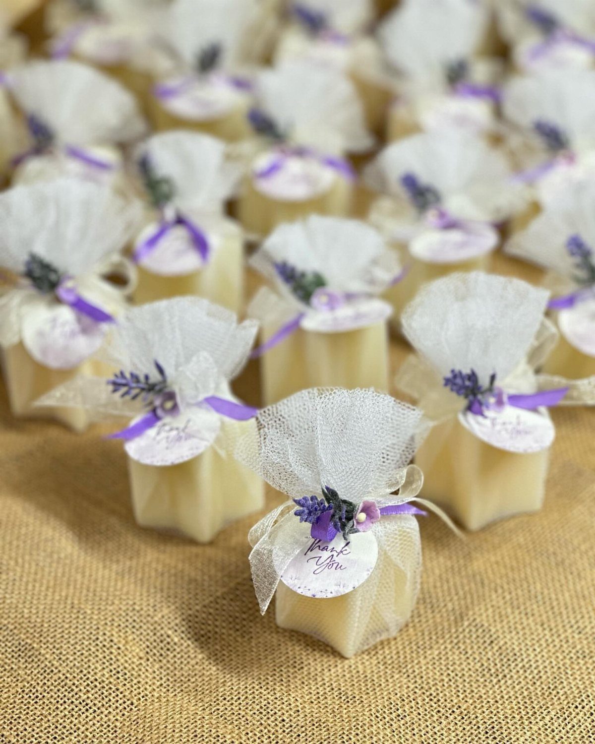 Small square cream candles with white tulle bows and lavender sprigs on burlap.