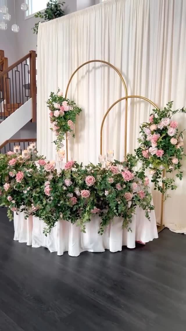 Gold metal hoop arches decorated with pink roses and greenery against white curtains.