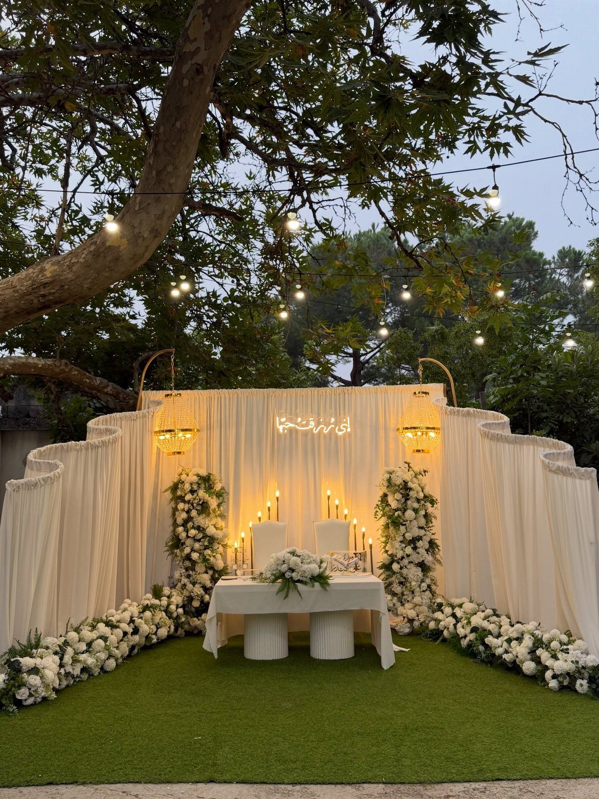 White fabric backdrop with floral arrangements and lanterns for an outdoor wedding ceremony.