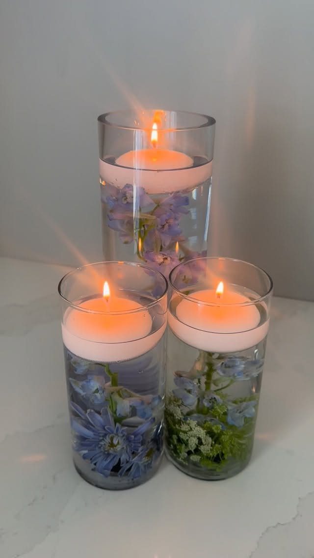 Three glass cylinder vases with floating candles and blue flowers in water.