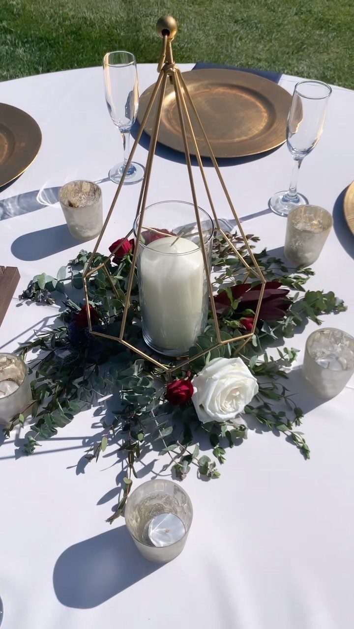 Gold geometric candle holder surrounded by eucalyptus greenery and roses on white tablecloth