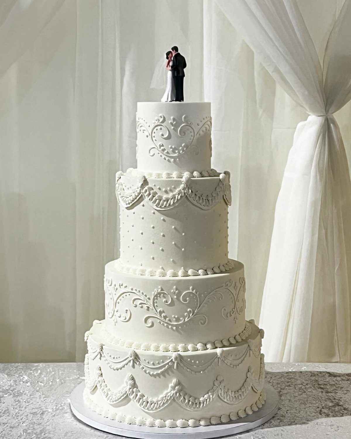 Four-tier white wedding cake with ornate frosting and a bride and groom topper.
