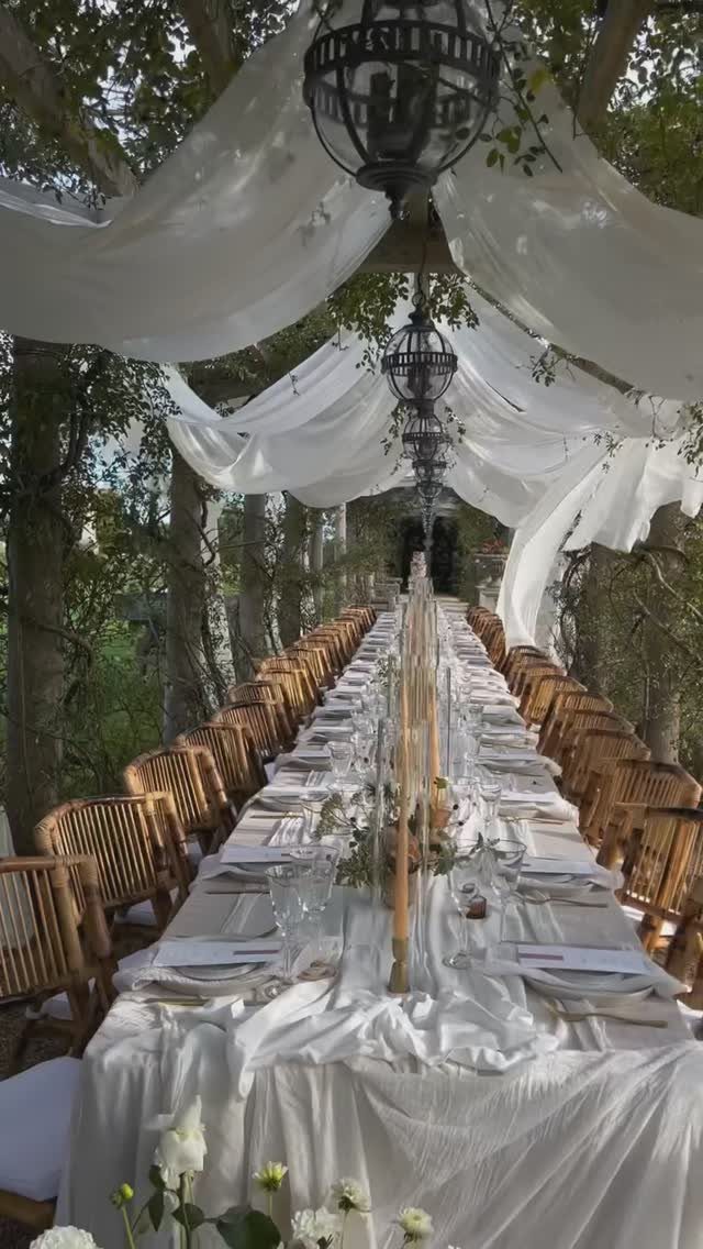 Long formal dining table with white linens and lanterns under draped white fabric.