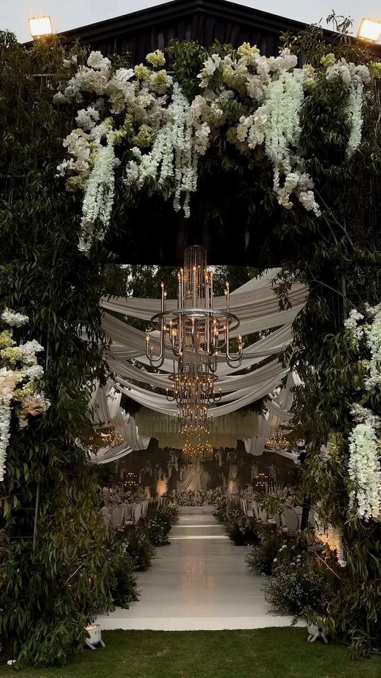 Floral arch framing an outdoor wedding venue with white drapes and crystal chandeliers.