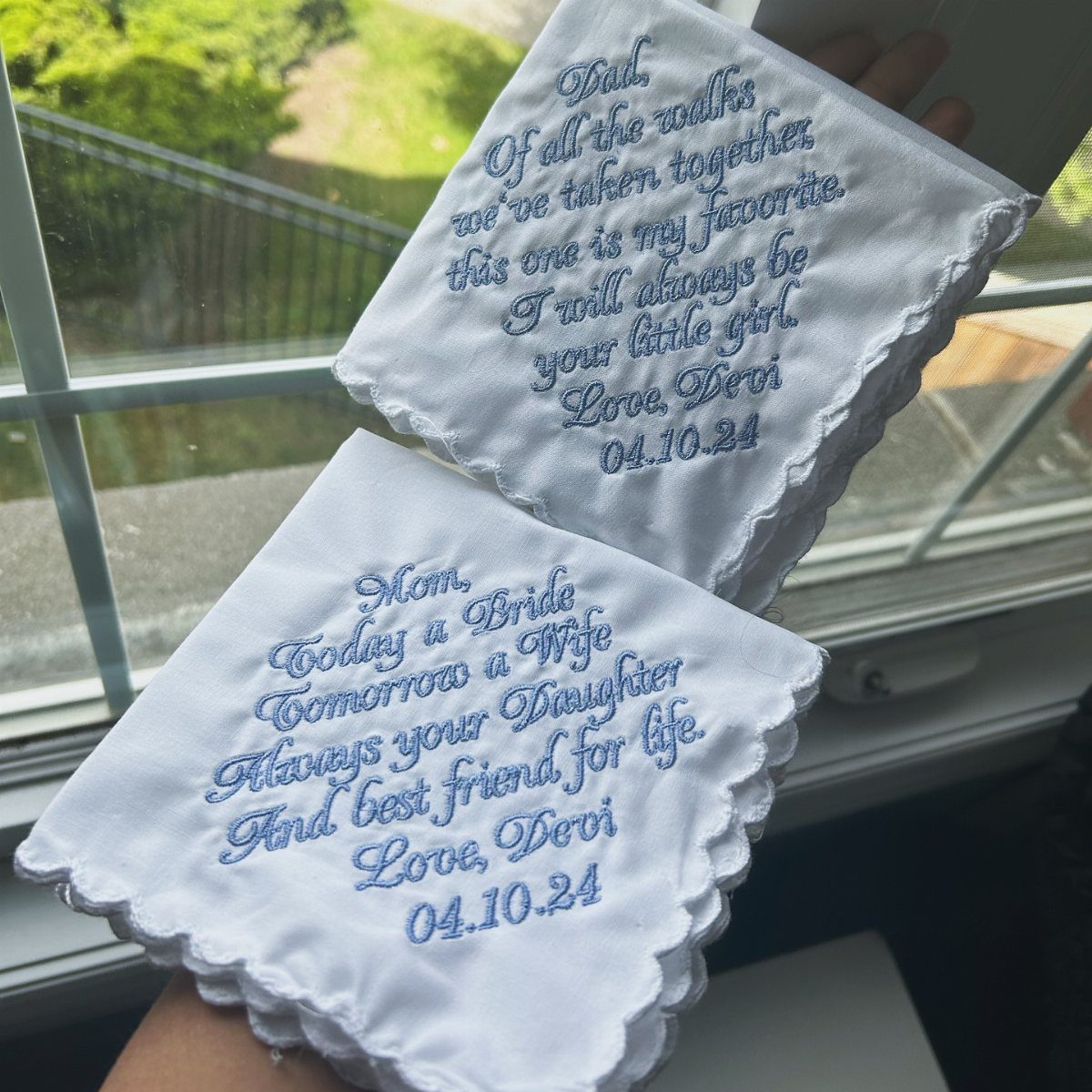 Two white embroidered handkerchiefs with blue text and scalloped edges held by hand.