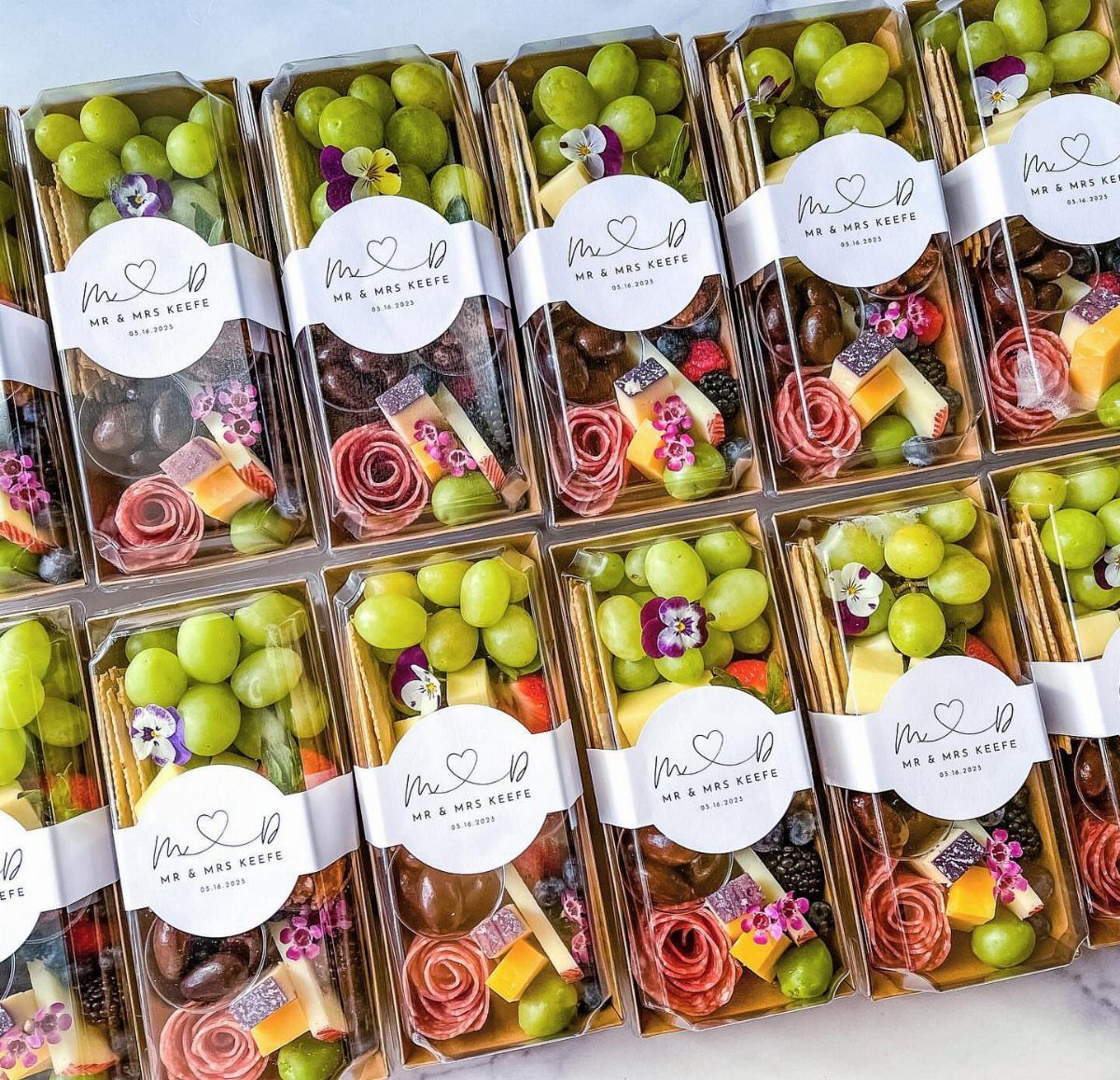 Assorted snack boxes with grapes, cheese, and meats for wedding guests.