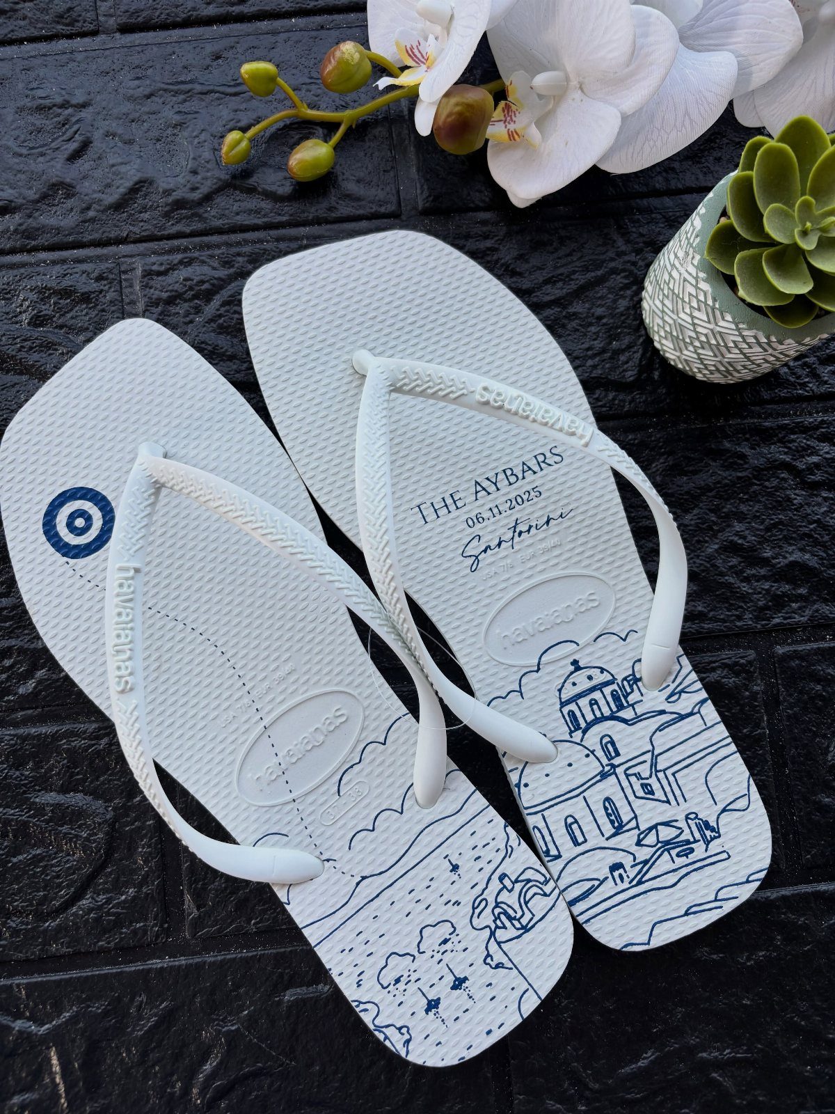 White personalized flip-flops with blue Greek scenery and evil eye illustrations on dark stone.