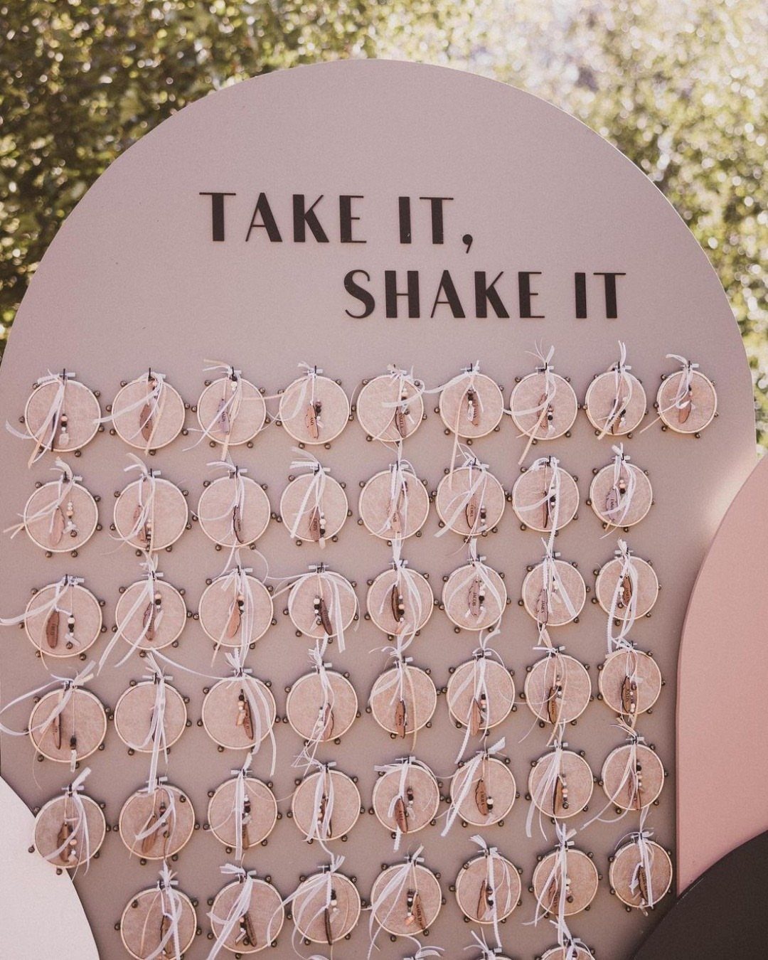 Wooden shaker bells attached to a pink arched board with wedding guest instructions.