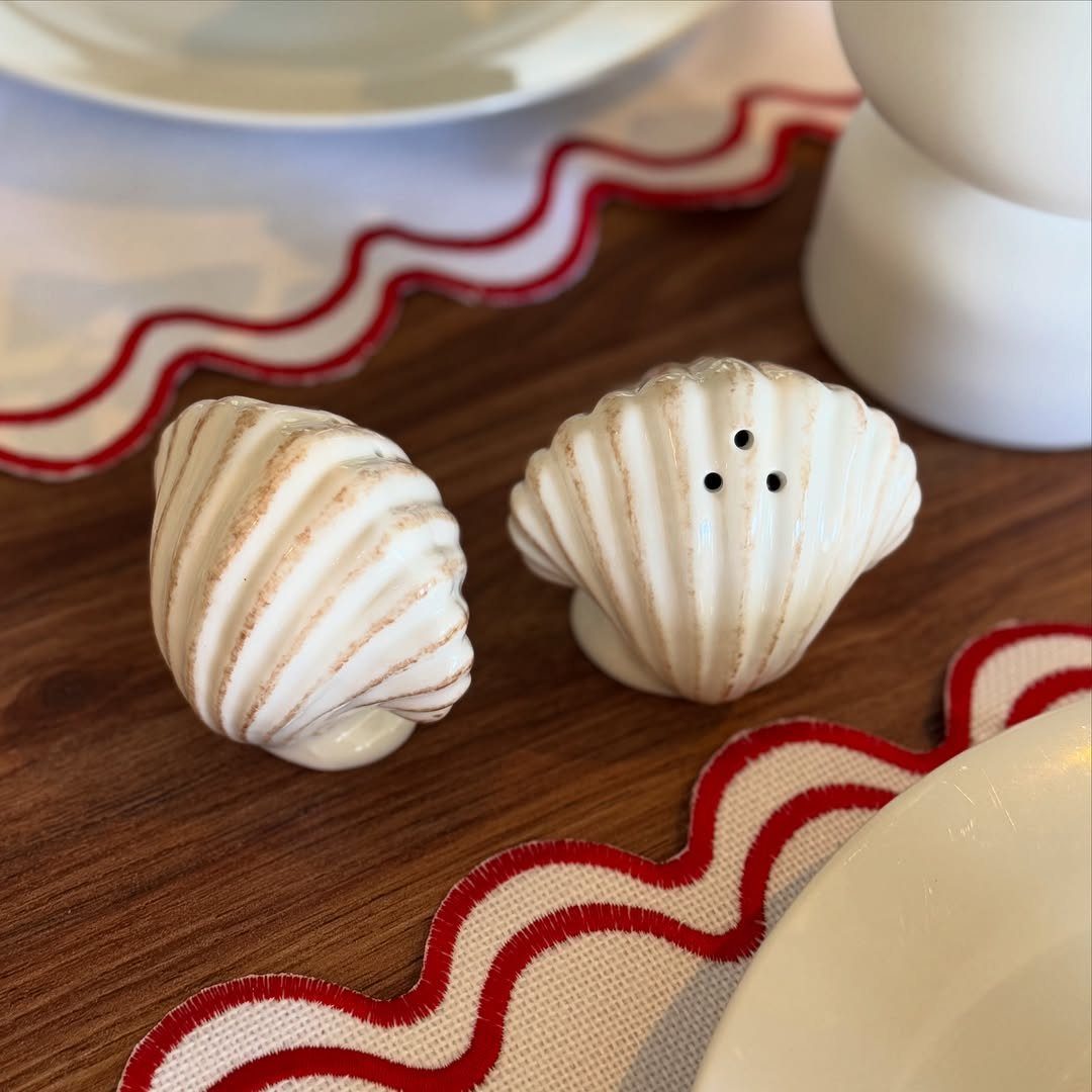 Seashell salt and pepper shakers on a wooden table with red scalloped placemats.