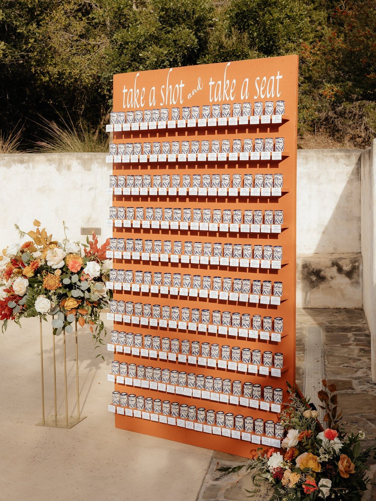 Orange wall display holding rows of shot glasses with name tags and floral arrangements.