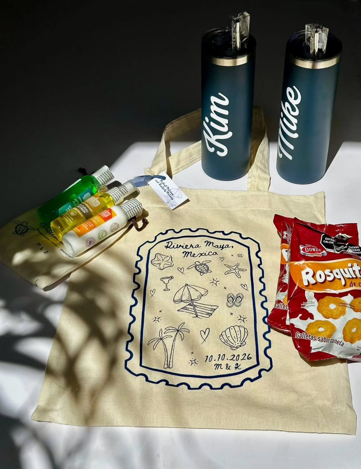 Canvas tote bag with beach illustration, personalized tumblers, and small sunscreen bottles.