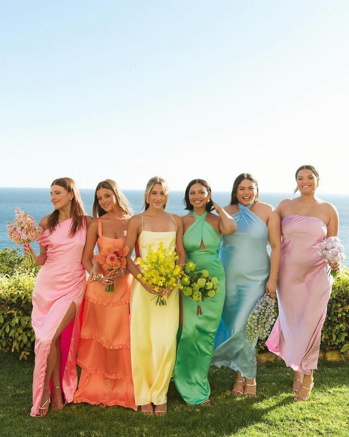 Six bridesmaids wearing different colored rainbow dresses standing on grass by the ocean