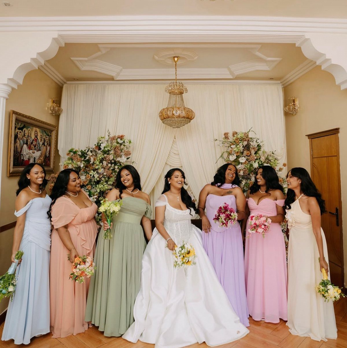 Bride in white gown surrounded by bridesmaids in various pastel colored dresses.