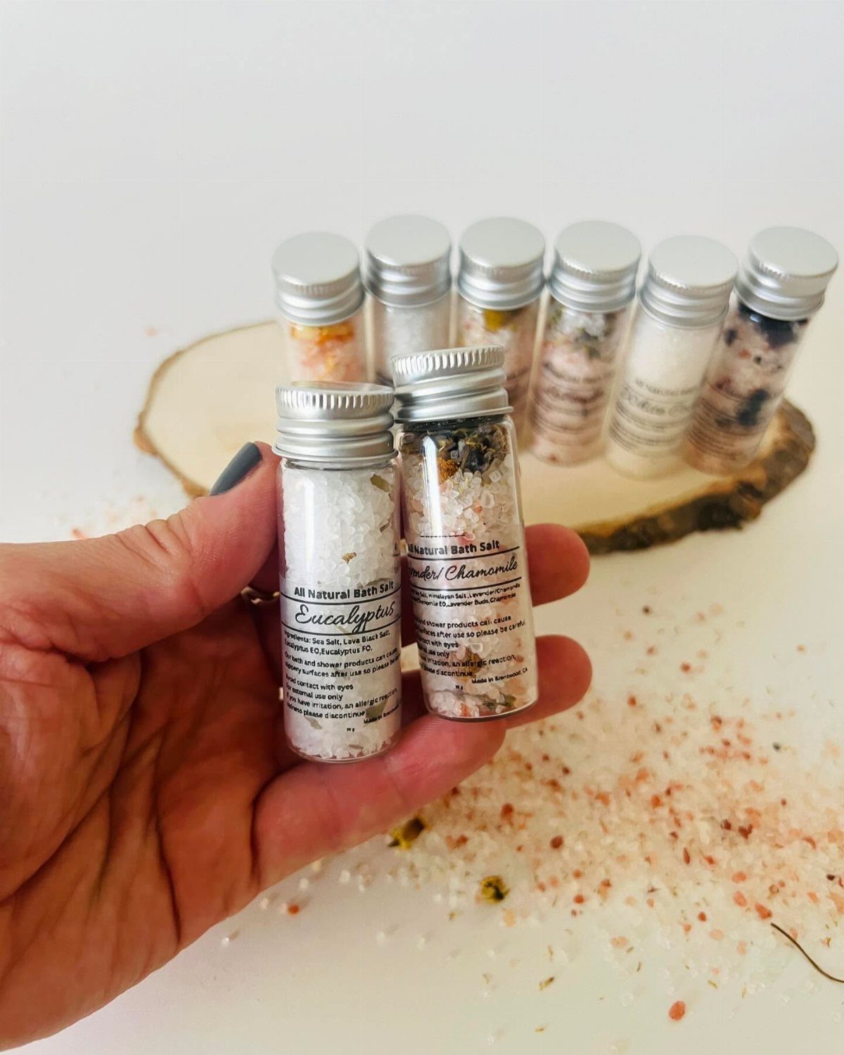 Small glass vials of colorful bath salts held in a hand and on wood.