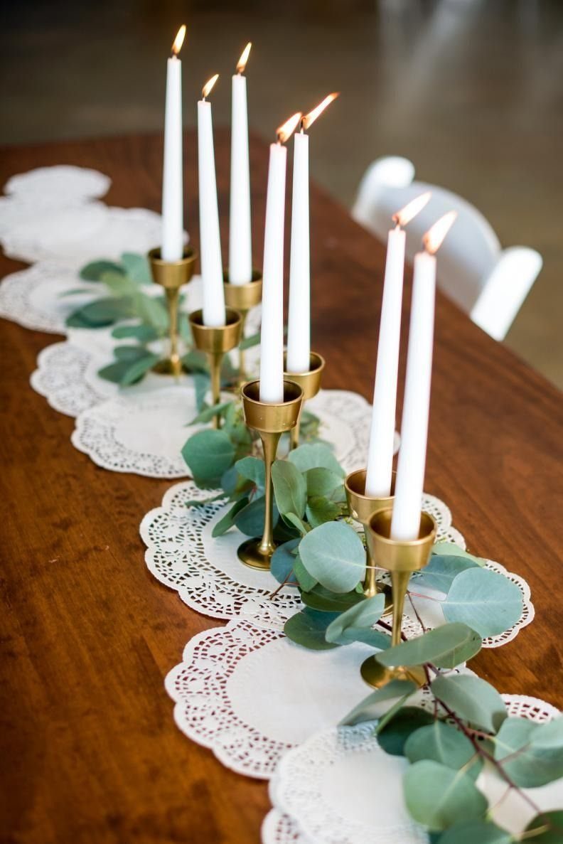 White taper candles in gold holders with eucalyptus leaves on lace doily runners.
