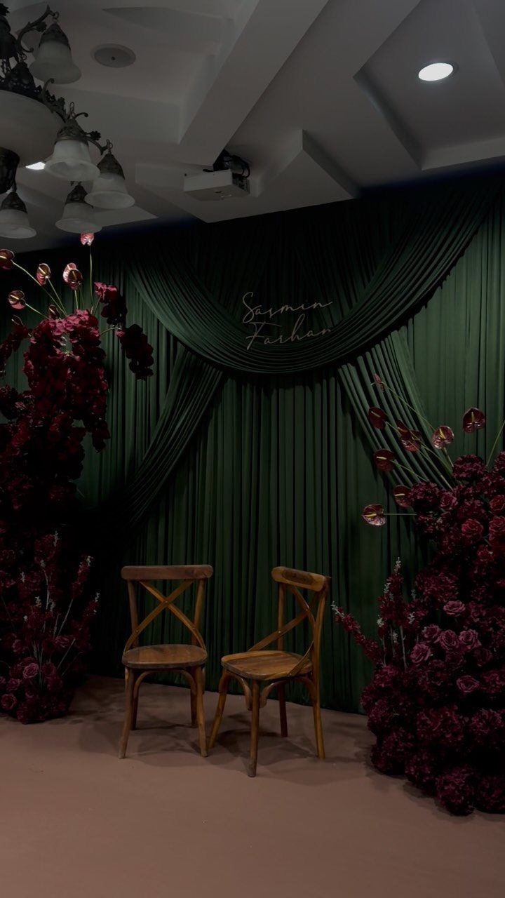 Two wooden chairs before an emerald green backdrop with burgundy floral arrangements.