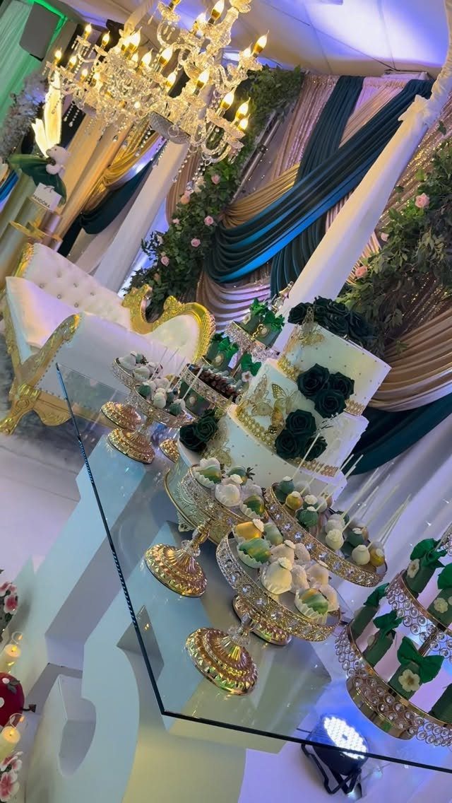 Dessert table with white cake, green roses, and gold stands against green drapery.